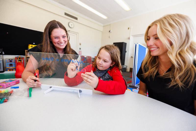 Two teaching aides assisting a child with their studies while smiling